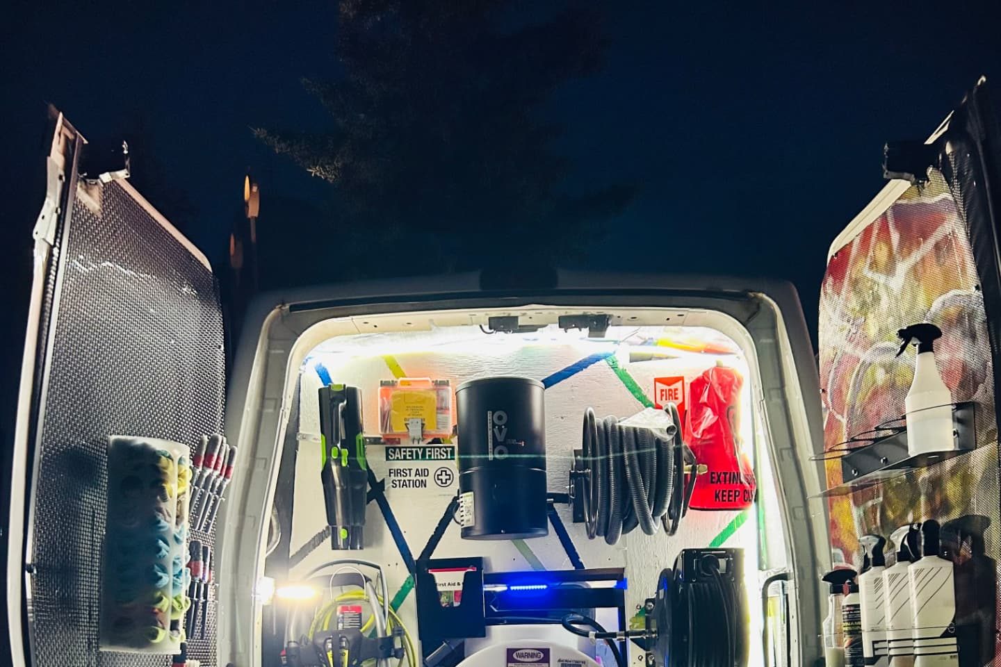 Rear doors of the SAKIGA CLEANS mobile detailing van open at night, showing an illuminated interior with a water tank, pressure washer, hose reels and organised detailing products